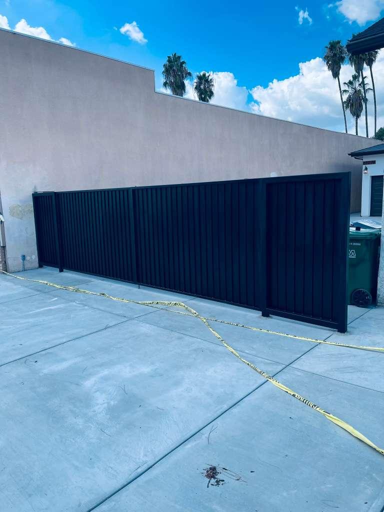 Modern black metal privacy fence with vertical panels installed along a residential driveway